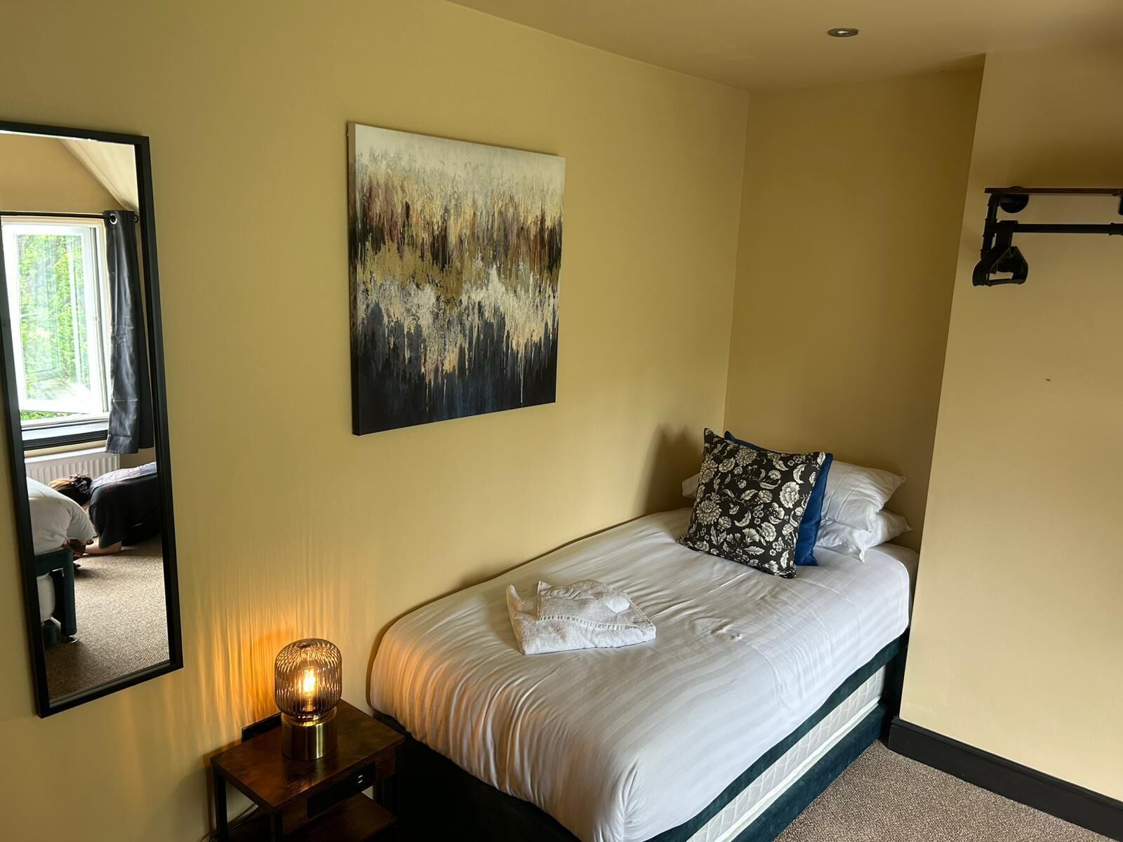 A hotel room with a bed, a bedside table with a lamp, a large abstract painting on the wall, and a window with curtains.