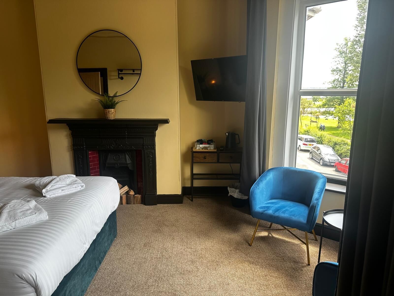 A hotel room with a bed, a blue armchair, a fireplace, a flat-screen TV, and a view of the outside.
