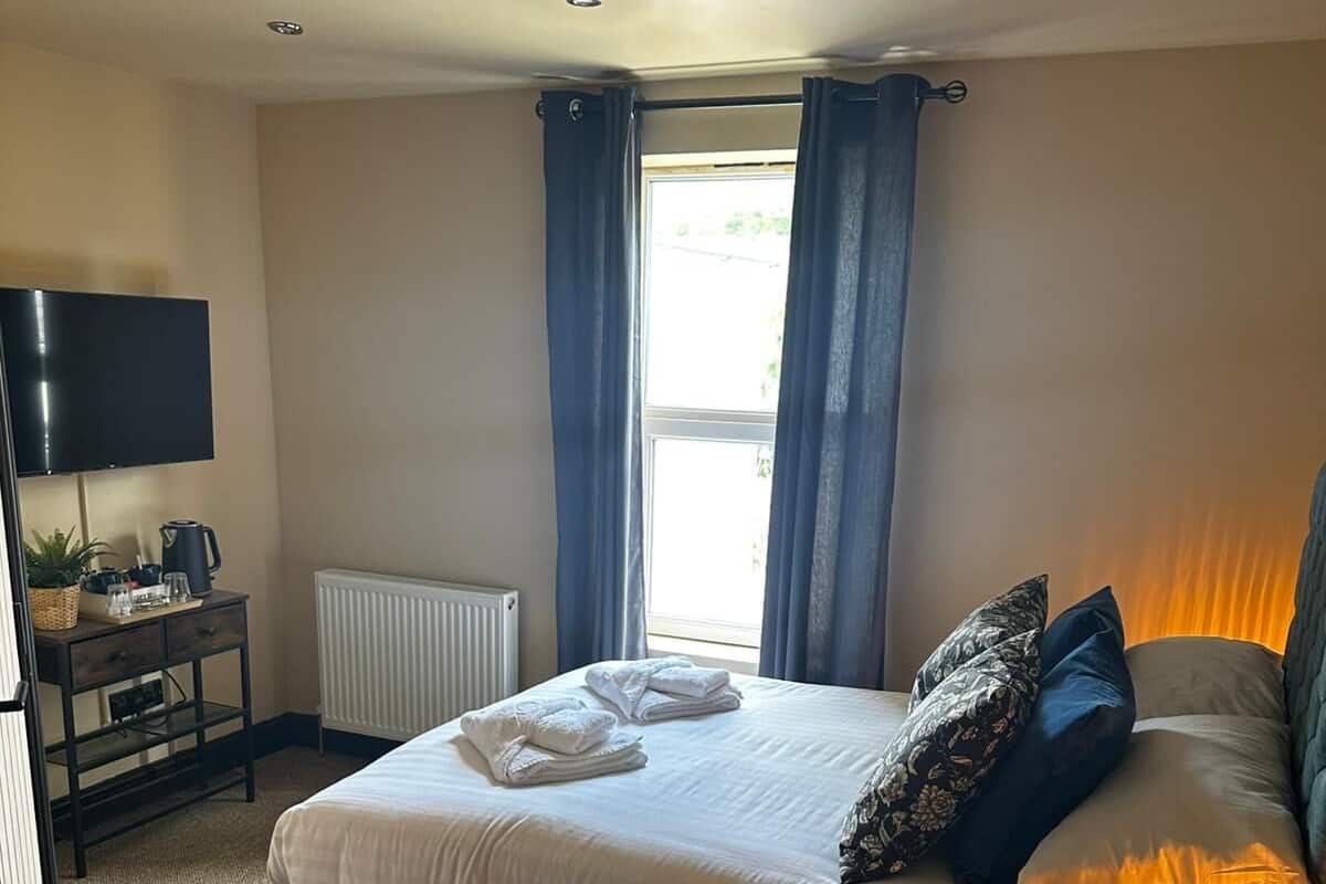A bedroom with a bed, a desk with a flat-screen TV, a radiator, and a window with curtains.
