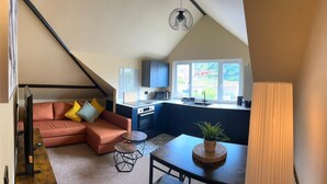 Un espacio habitable compacto con kitchenette, un sofá con cojines, una pequeña mesa de comedor y una planta en maceta.