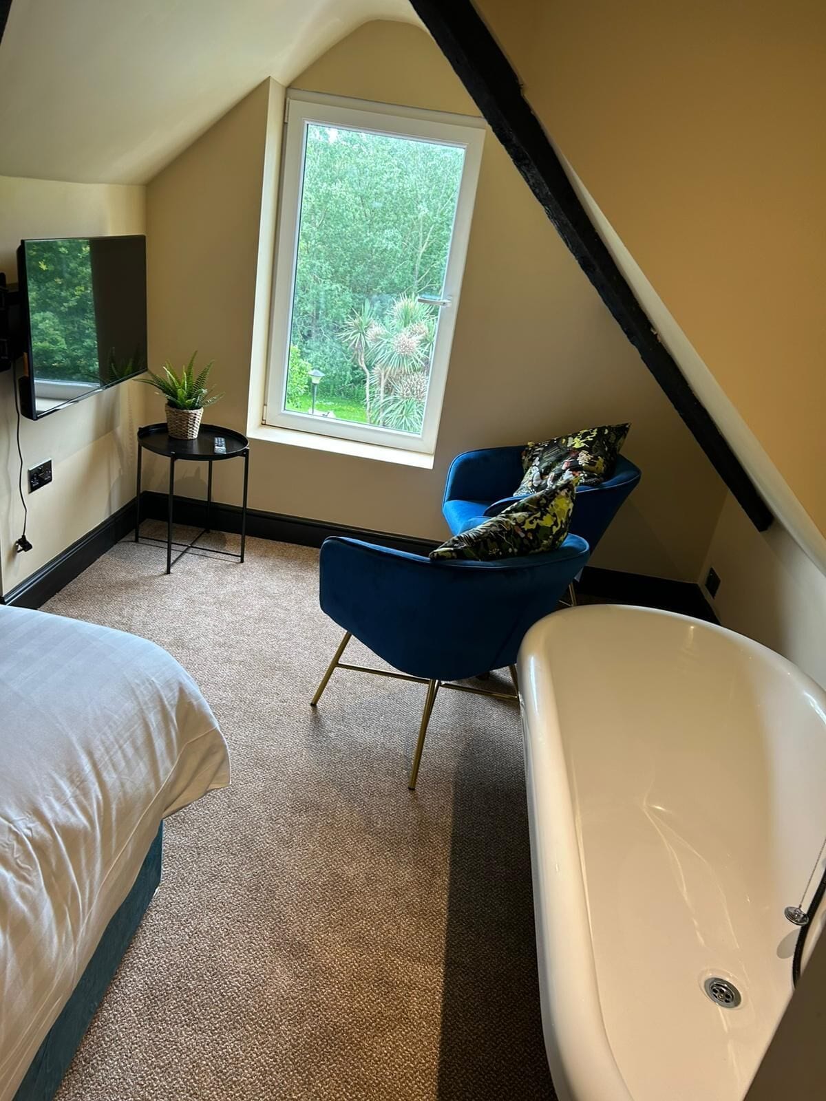 Un dormitorio con un ventanal grande, una silla azul, una bañera blanca y una cama.