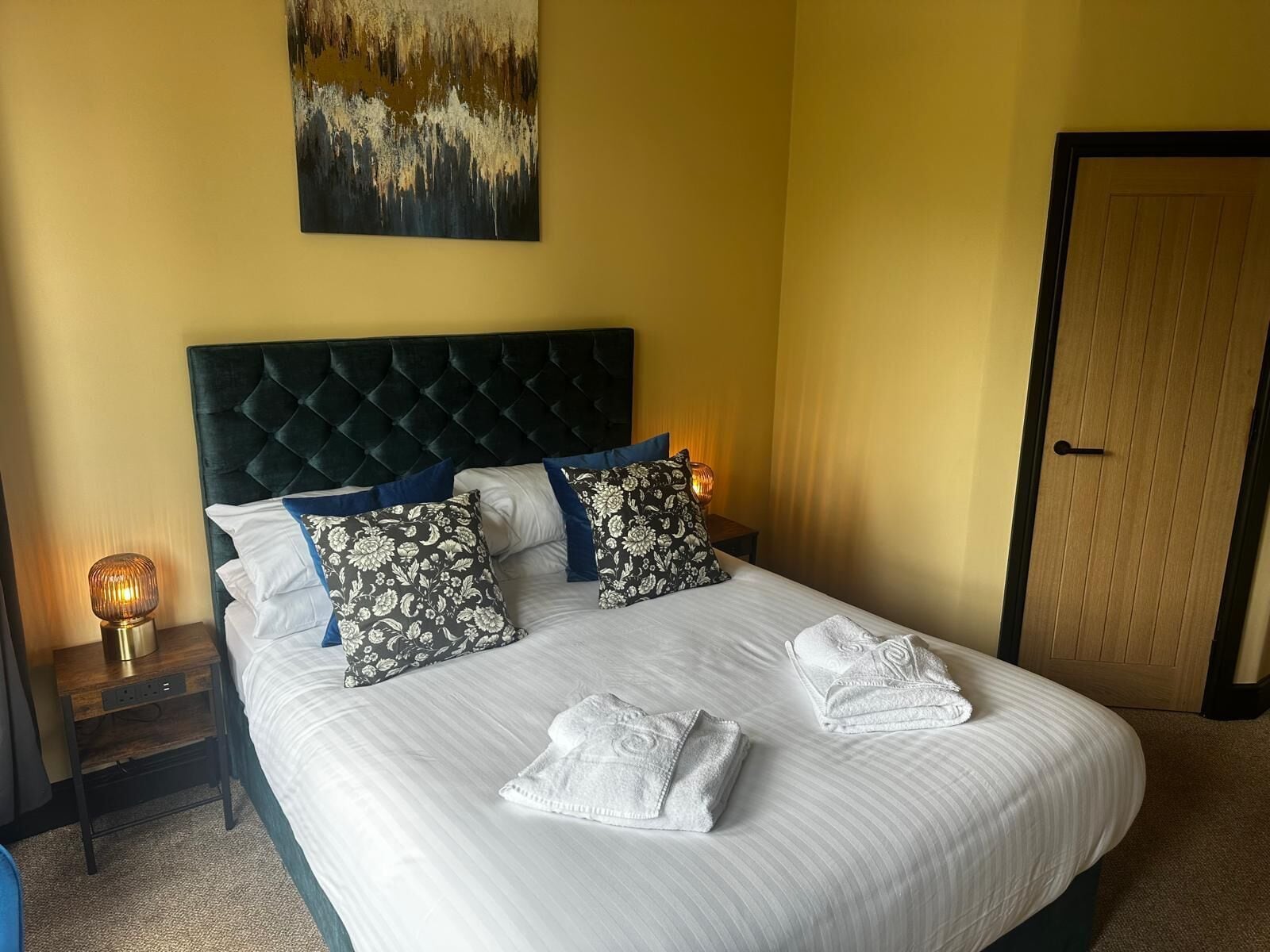A neatly made bed with white linens and two decorative pillows, a bedside lamp, and a painting on the wall.