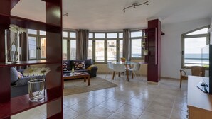 A spacious living area with a sofa, coffee table, and dining table. Large windows offer a view of the sea.