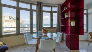 A room with a glass table, white chairs, a red bookshelf, and a view of the sea through large windows.