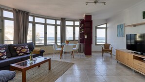 A modern living room with a sofa, coffee table, and TV stand. There is a dining area with a table and chairs. Large windows offer a view of the sea.