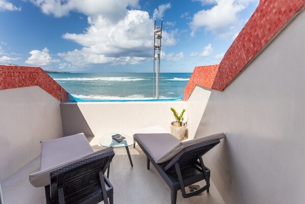 A balcony with two wicker chairs, a small table, and a potted plant. The balcony overlooks the ocean with waves crashing against the shore.