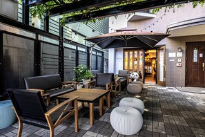 An outdoor seating area with wooden tables and chairs, a large umbrella, and a brick wall in the background. - KIZEN VILLA (Tokyo)