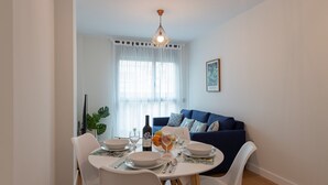 Een eetgedeelte met een ronde tafel voor vier personen, witte stoelen, een blauwe bank op de achtergrond en een groot raam met dunne gordijnen.