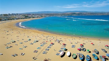 A beach with rows of umbrellas and boats, a clear blue sea, and a distant cityscape.