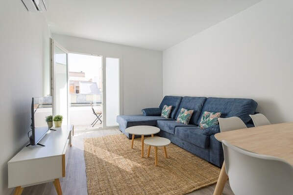 Ein modernes Wohnzimmer mit einem blauen Sofa, einem kleinen weißen Tisch, einem Fernseher auf einem Lowboard und einem großen Fenster.