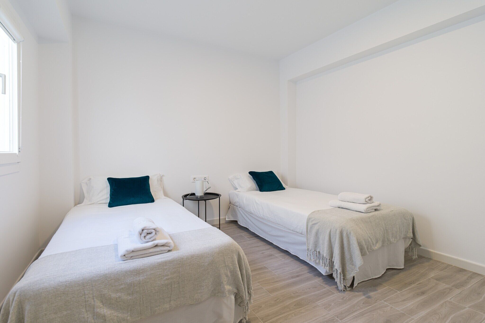 A clean, minimalist hotel room with two single beds, a small bedside table, and a window.