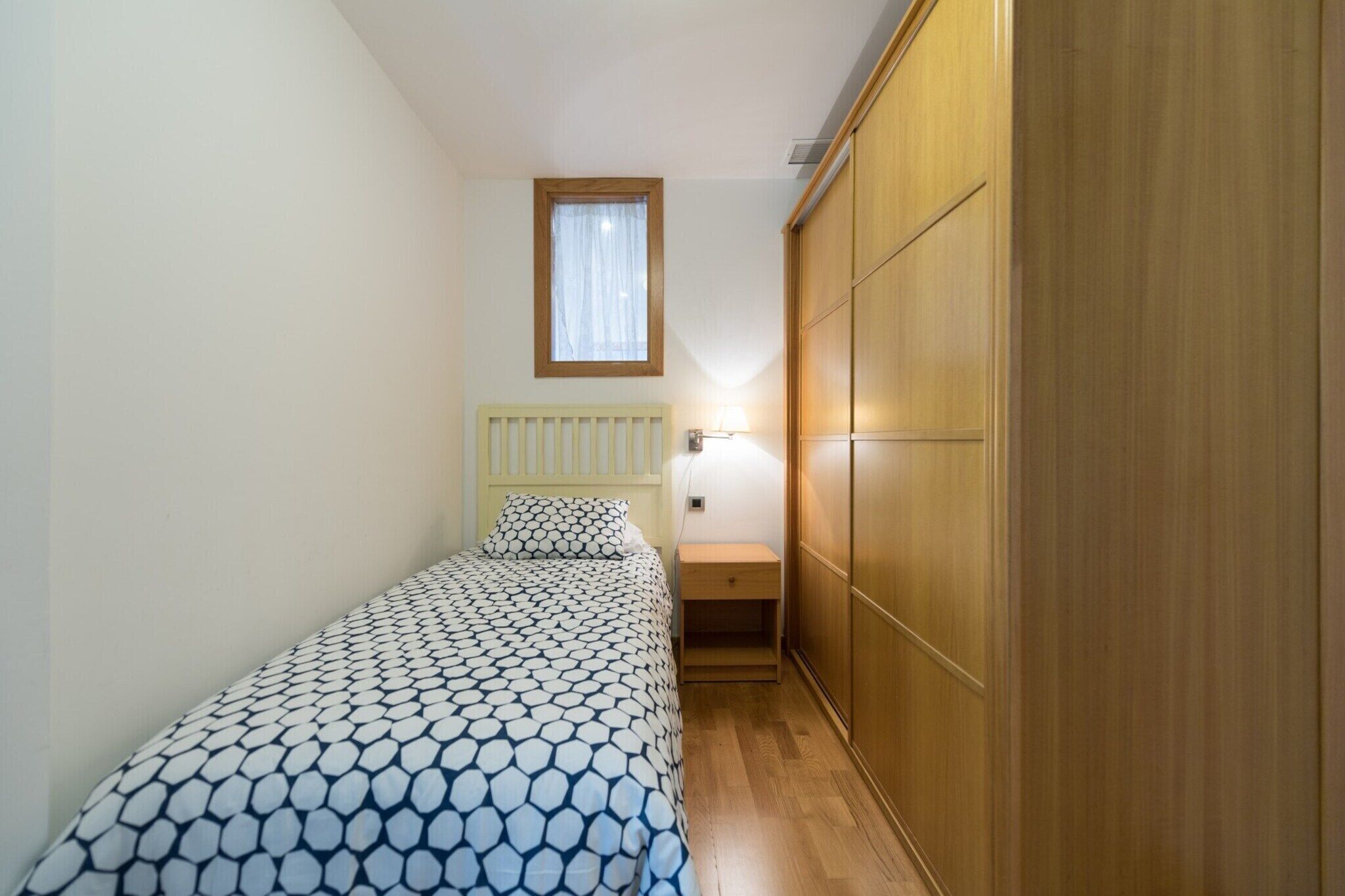 A small bedroom with a bed, a nightstand, a wooden wardrobe, and a window with a curtain.