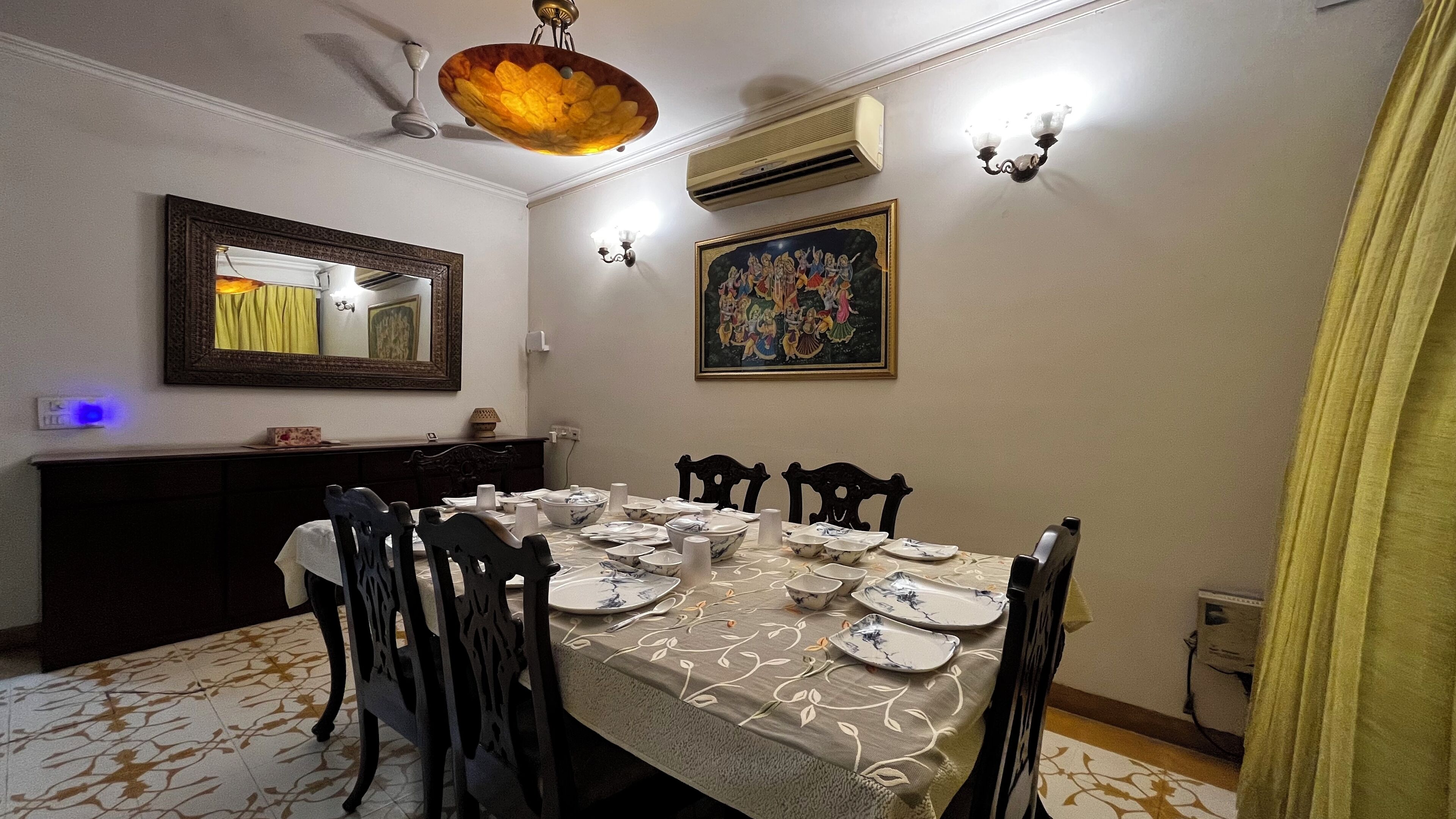 Un coin repas avec une table dressée pour quatre personnes, entourée de chaises. On y trouve un ventilateur de plafond, un appareil de climatisation et des luminaires fixés au mur.