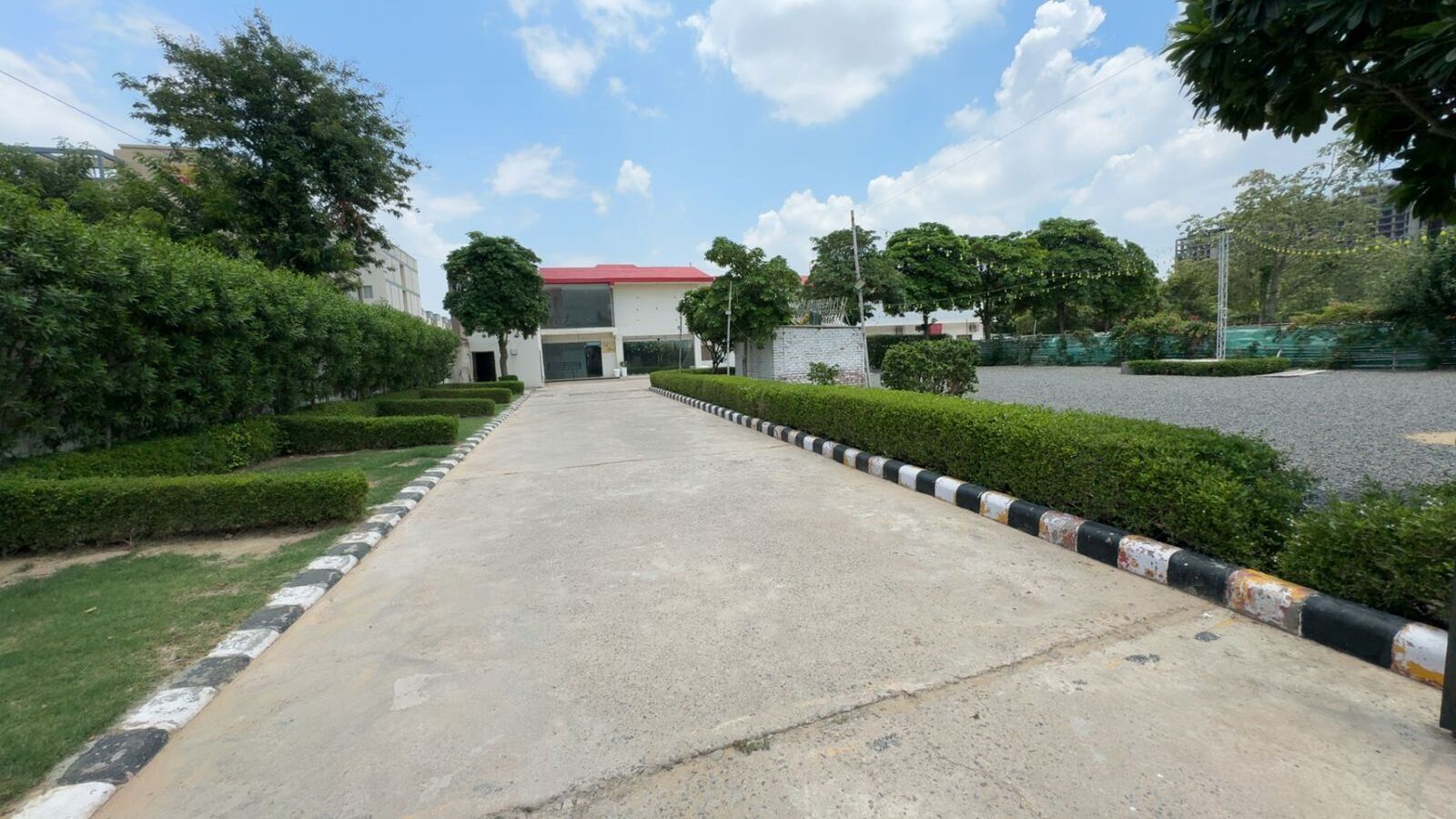 A paved pathway leading to a building with a red roof, flanked by neatly trimmed hedges and trees.