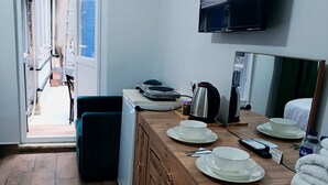 Ein kleines Hotelzimmer mit einem Holztisch für vier Personen, einem kleinen Kühlschrank, einem an der Wand montierten Fernseher und Blick auf den Außenbereich des Ganges.
