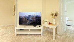 Una sala de estar con un televisor de pantalla plana sobre un pedestal blanco, una mesita auxiliar blanca con un florero y objetos decorativos, y una puerta que da a otra habitación.