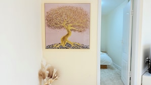 Una habitación con un cuadro de un árbol en la pared, un jarrón blanco con plantas secas y una puerta que da a otra estancia.