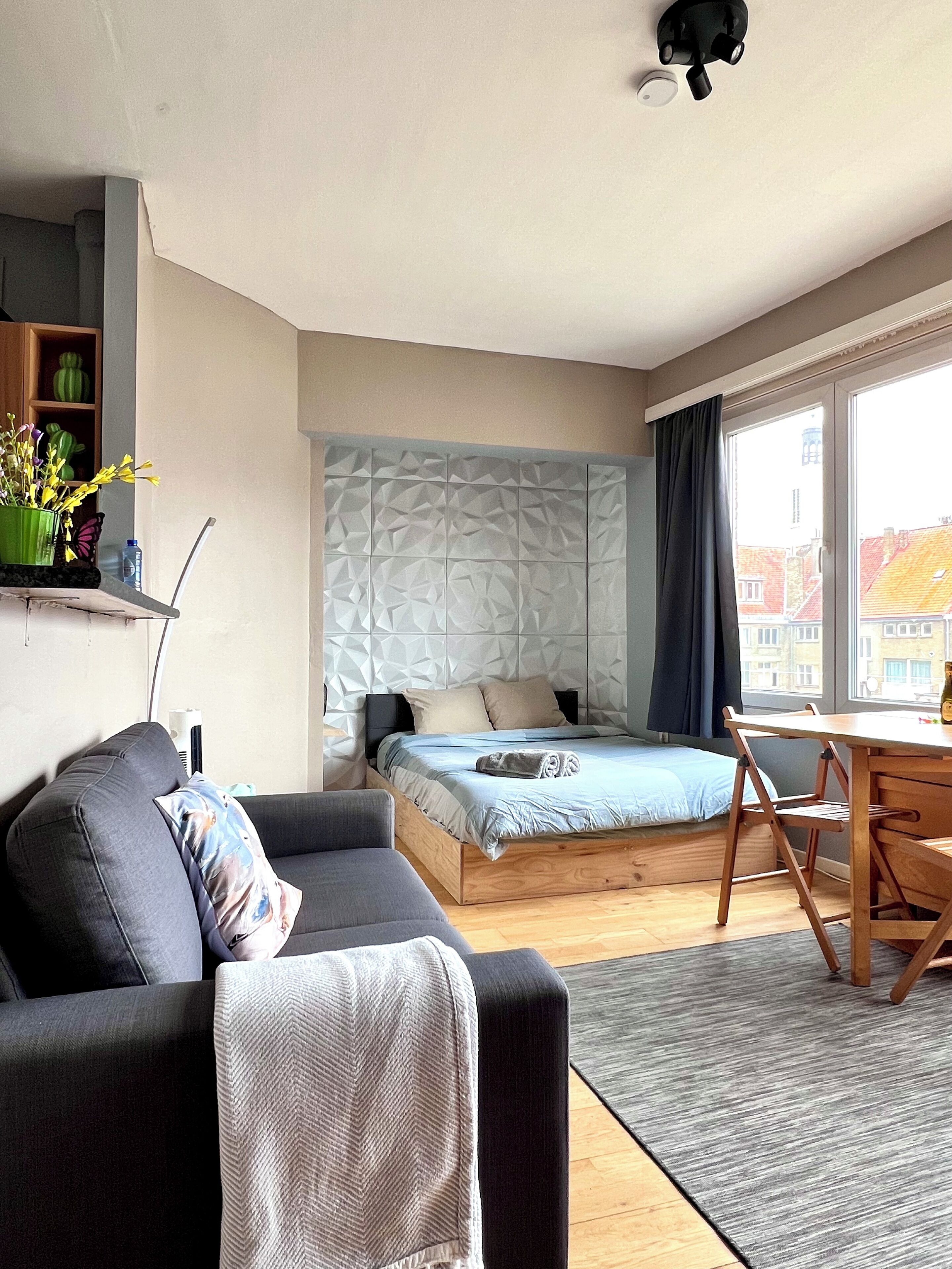 A modern bedroom with a bed, a sofa, a desk, and a window with a view of buildings.