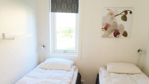 Ein kleiner, sauberer Raum mit zwei Einzelbetten, einem Fenster mit einem gemusterten Rollo und einem Blumenbild an der Wand.