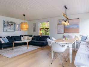 A modern living room with a dark-colored sofa set, a wooden coffee table, and a white dining table with chairs. There are framed pictures on the walls, a hanging light fixture, and a view of greenery outside. - 4 Star Holiday Home in Albaek (Aalbaek)