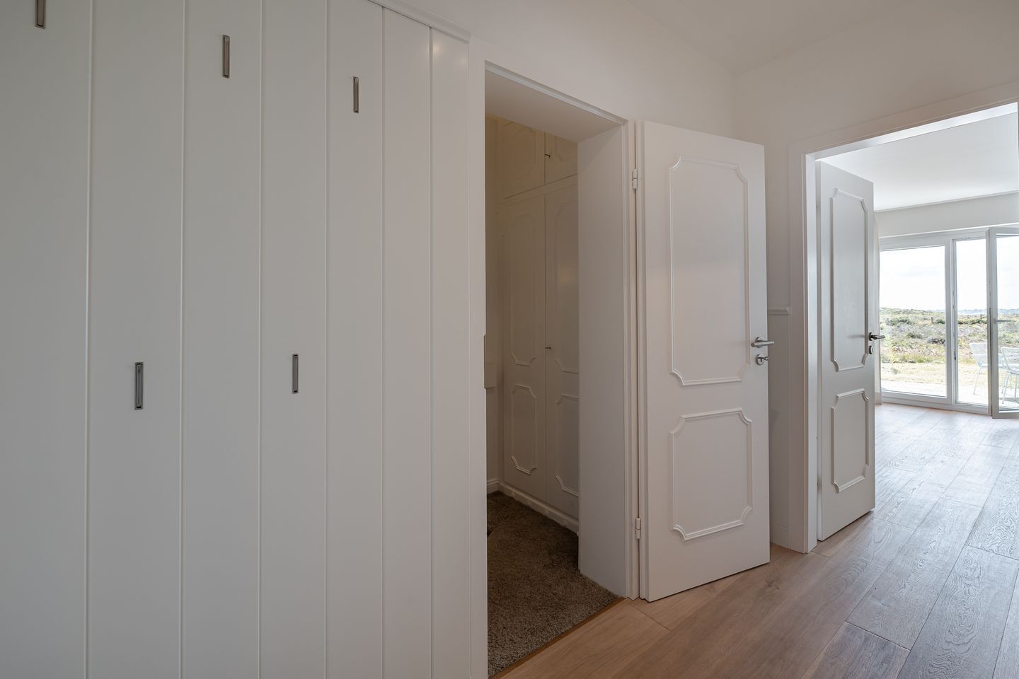 Un couloir avec des portes blanches, un espace recouvert de moquette et une vue à travers une porte vitrée.