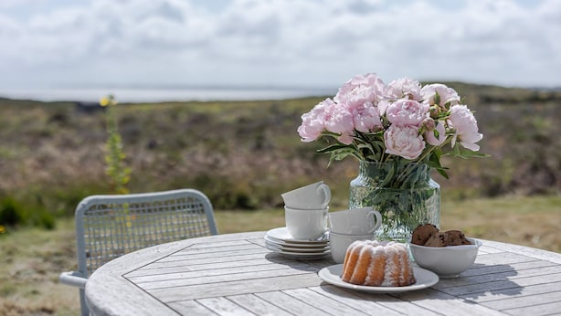 A round wooden table with a white vase of pink flowers, a plate with a bundt cake, and a bowl of cookies.