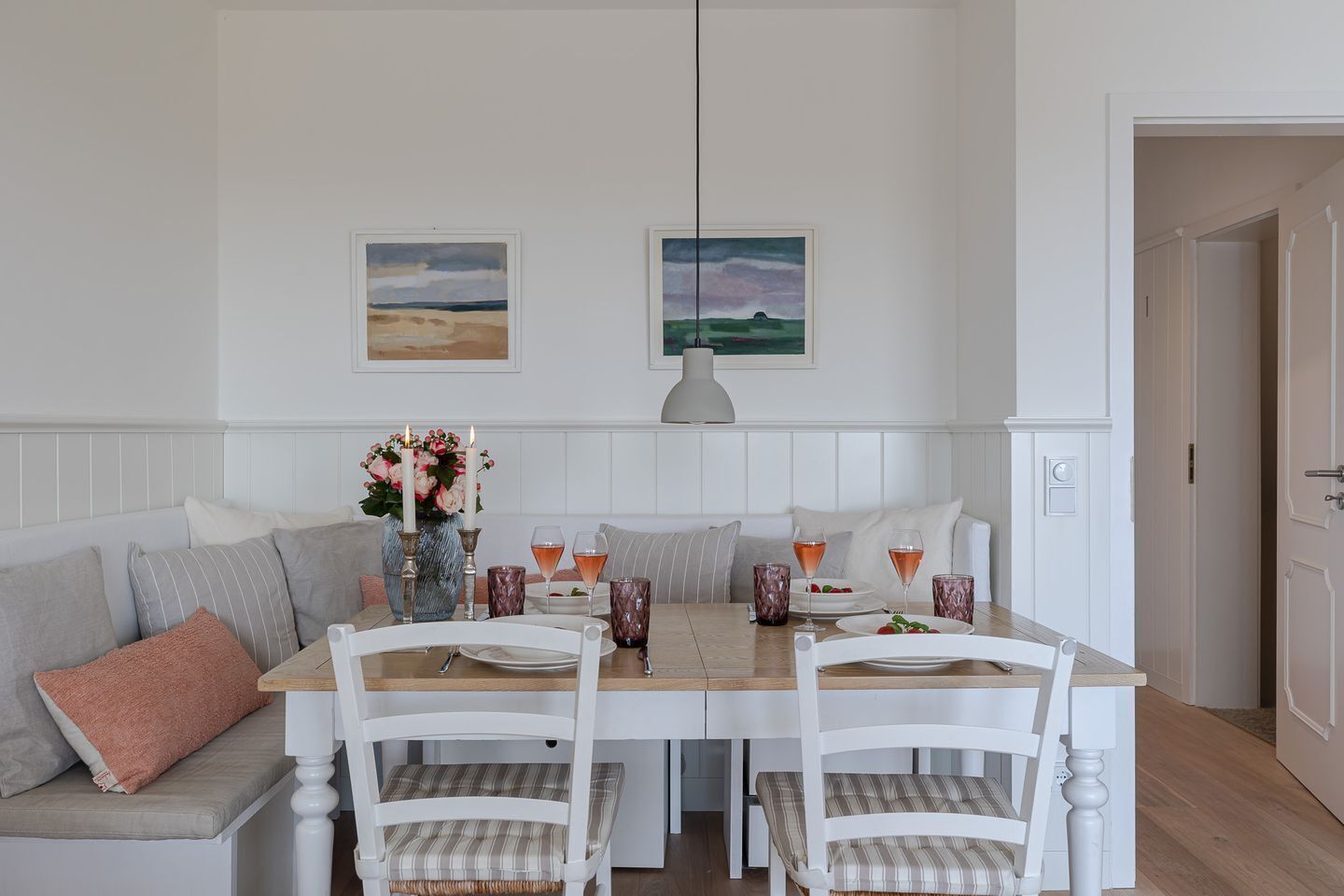 A dining area with a wooden table, white chairs, a bench, and two paintings on the wall.