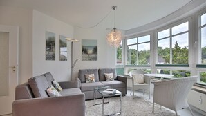 Un salon moderne avec un canapé gris, une table basse en verre et un fauteuil en osier. De grandes fenêtres offrent une vue sur la verdure environnante.