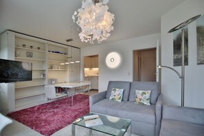 Un salon moderne avec un canapé gris, une table basse en verre, un tapis à motifs floraux rouges, une bibliothèque blanche, un téléviseur à écran plat et un lustre blanc original.