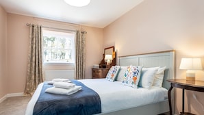 A neatly made bed with pillows, a bedside table with a lamp, a window with curtains, and a radiator. - Byre Cottage | Stravithie (Stravithie)