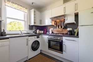A modern kitchen with white cabinets, a washing machine, an oven, and a window with a view of trees. - Kidston Court (No 9) (St Andrews)