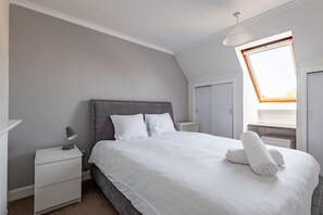 A neatly arranged bedroom with a large bed, a bedside lamp, a nightstand, and a window with a view. - Kidston Court (No 9) (St Andrews)