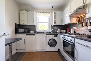 A compact kitchen with white cabinets, a washing machine, a microwave, and a stove. - Kidston Court (No 9) (St Andrews)