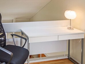 Un escritorio blanco con una lámpara, una silla de oficina negra y una ventana con persianas.