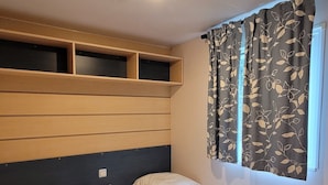 A room with two single beds, a wooden headboard, a bedside table, and a window with floral curtains. - MobileHome VERONA, air-conditioned, TV, WIFI, 15 min from Europa-Park! (Boofzheim)