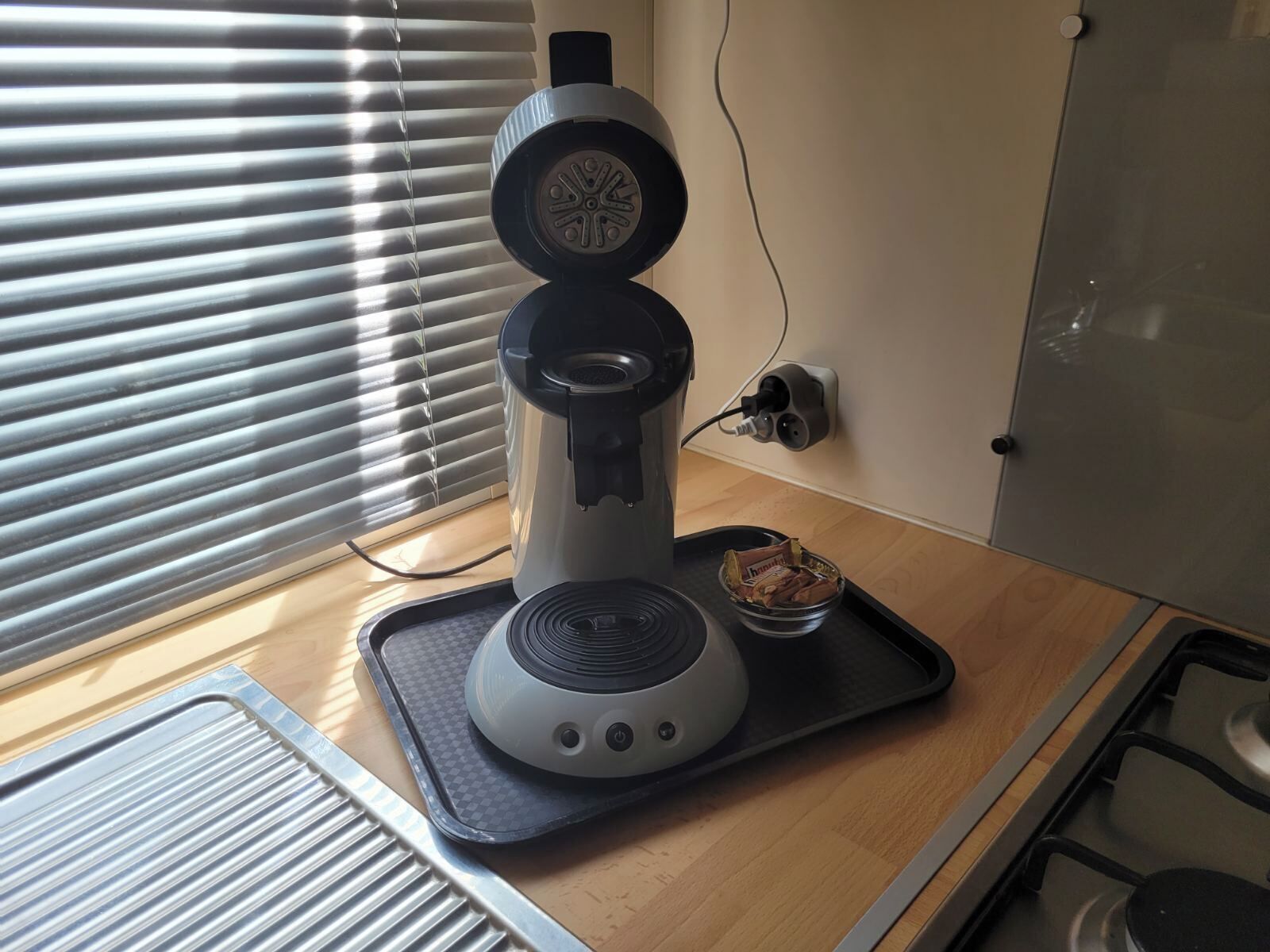 Une cuisine avec une machine à café posée sur le plan de travail, une fenêtre avec des stores et un petit bol de collations.