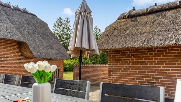Un patio con una mesa lista para una comida, un florero con flores blancas y una cesta de frutas.