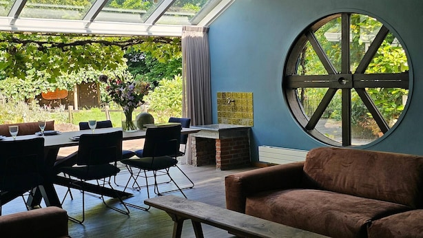 A dining area with a table, chairs, and a bench. There is a large window with a view of greenery outside.