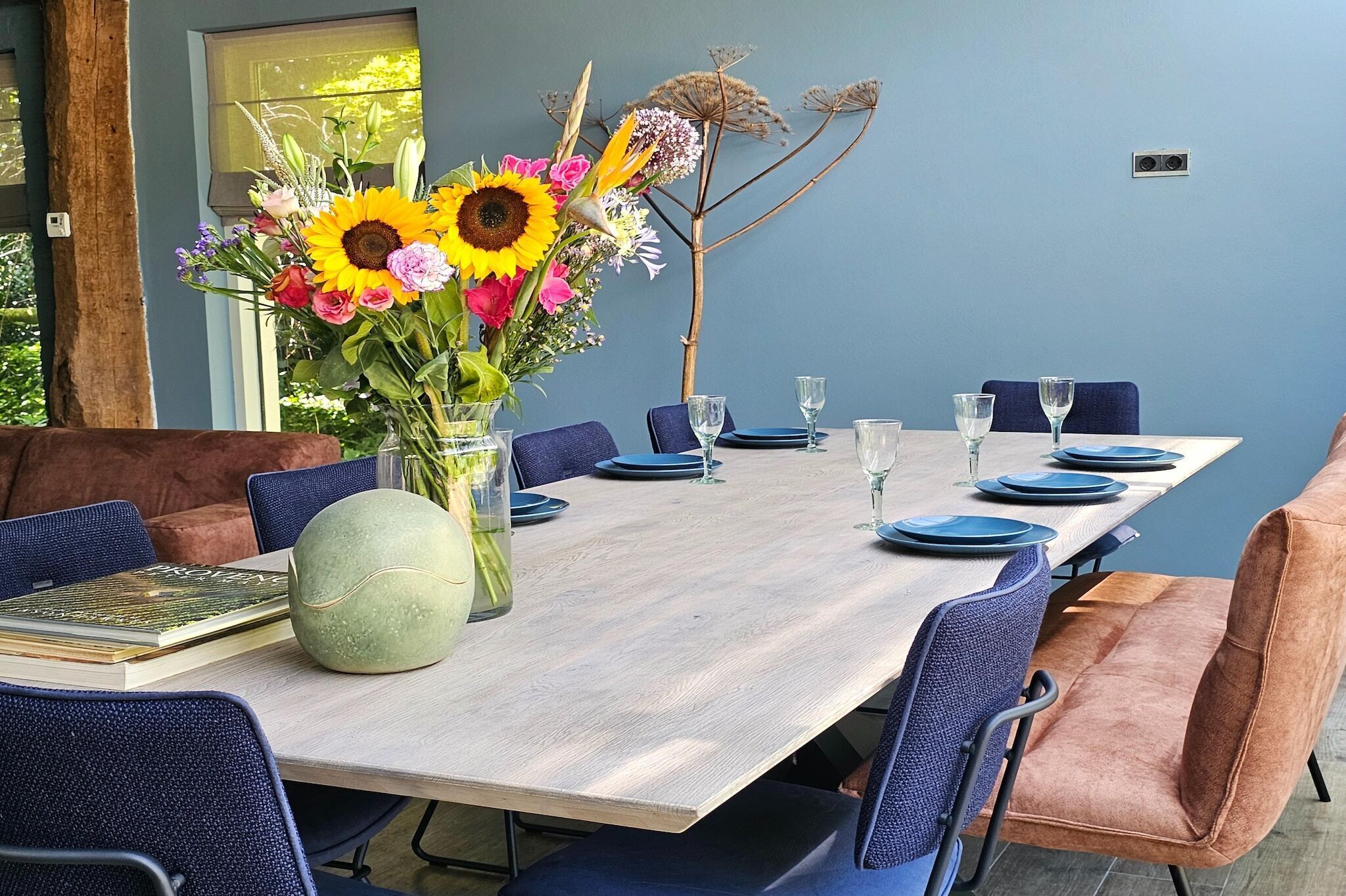 Un coin repas avec une longue table dressée pour six personnes, des chaises bleues et un vase rempli de fleurs.
