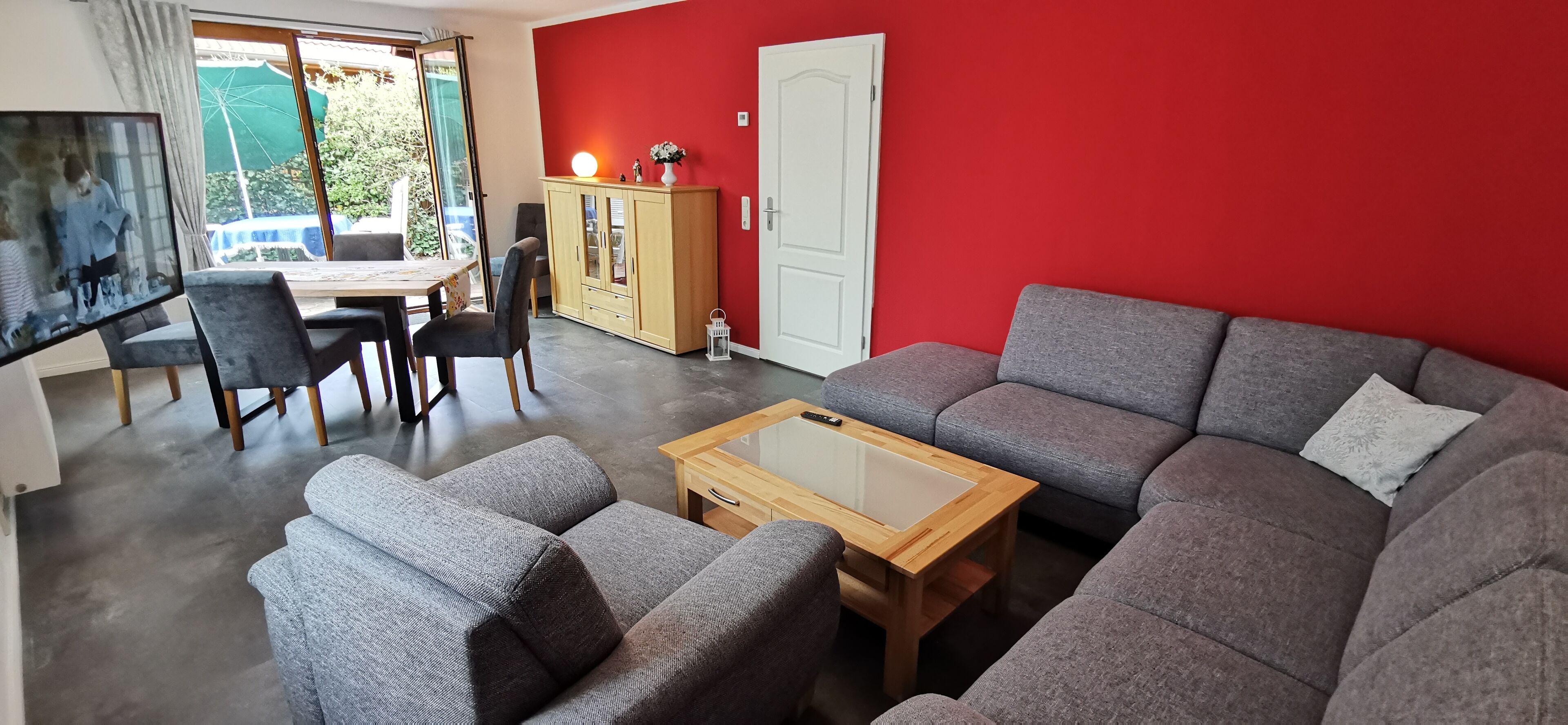 A living room with a red wall, grey sofa, wooden coffee table, and a dining area with a table and chairs.