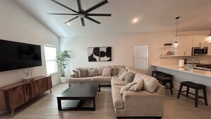 A modern living room with a sectional sofa, a flat-screen TV, a ceiling fan, and a kitchen area with a microwave.