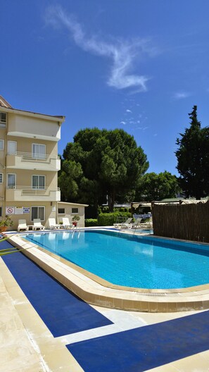 A swimming pool with lounge chairs, a building with balconies, and trees in the background.