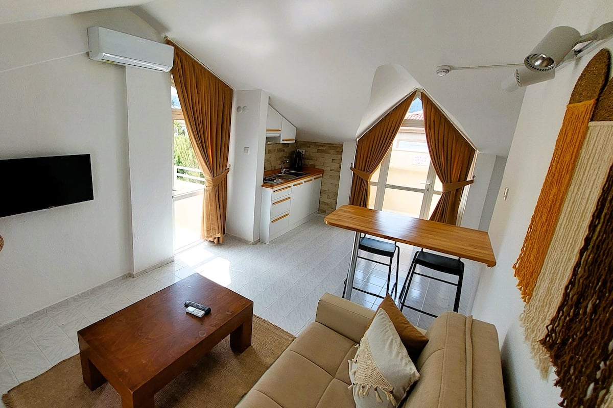 Een knus leefruimte met een aan de muur bevestigde televisie, een houten salontafel, een bank en een keukenhoek met een raam.