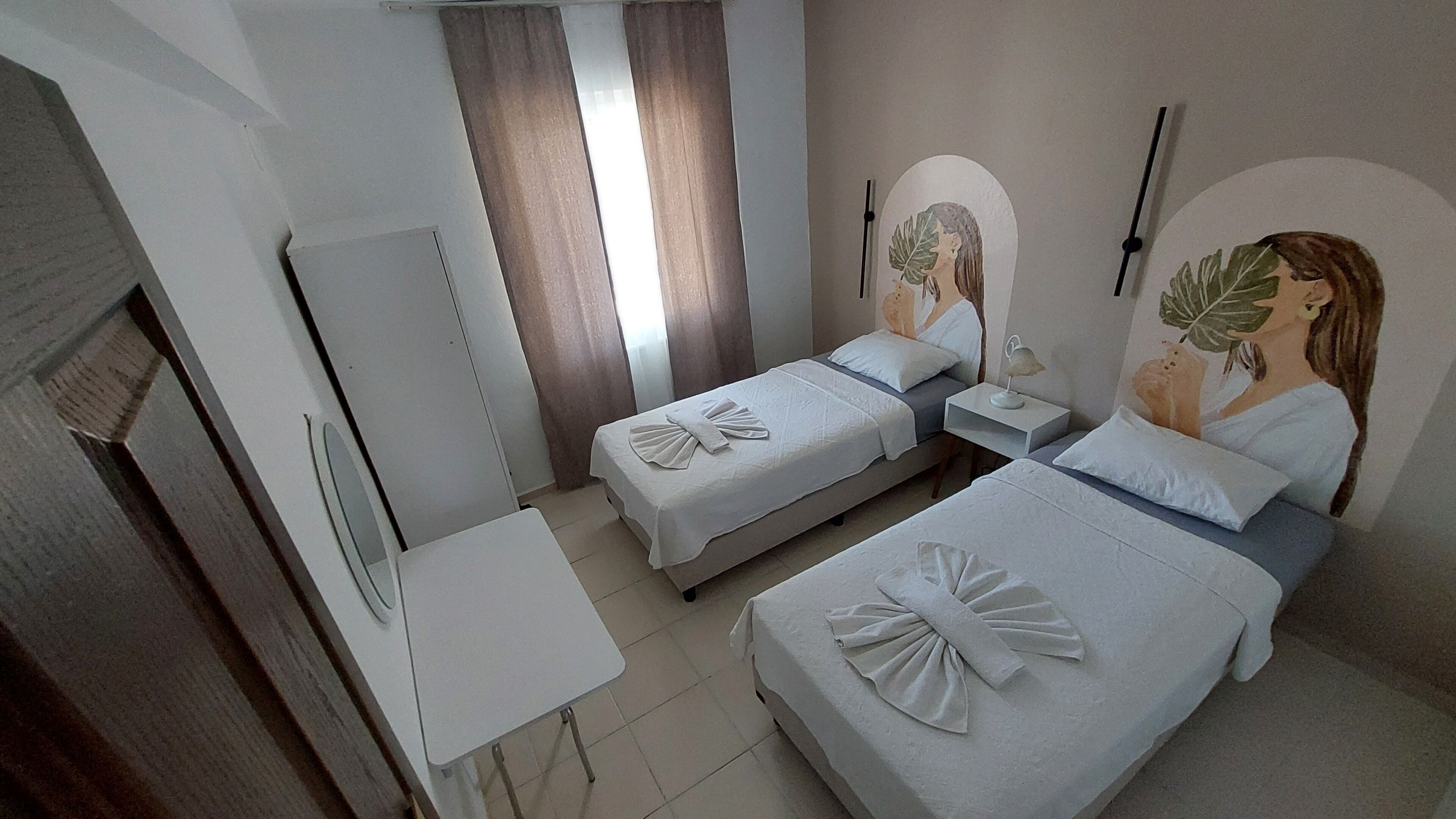 A hotel room with two single beds, a small table, a mirror, and a wall-mounted lamp.