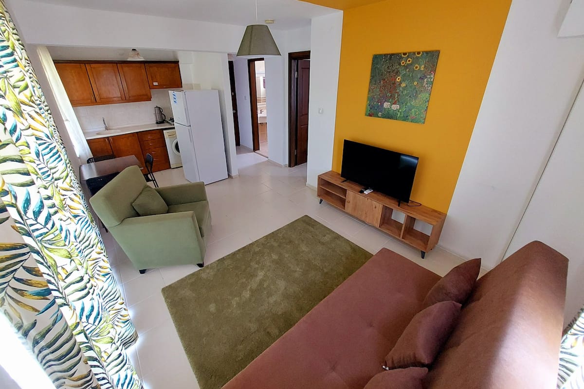 Een woonkamer met een bank, een televisie op een houten standaard, een keuken op de achtergrond, en een kleurrijk schilderij aan de muur.