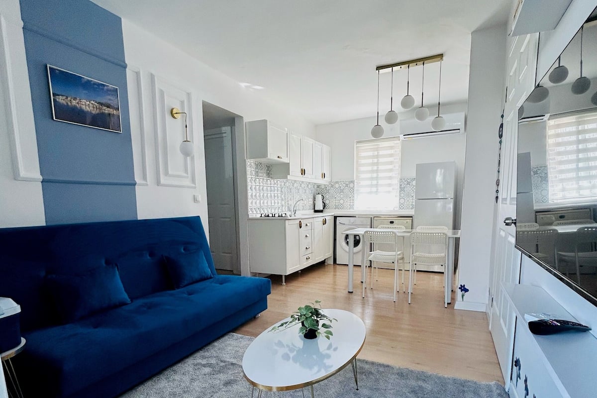 Een moderne woonkamer met een blauwe bank, een klein rond salontafeltje, en een keuken op de achtergrond.