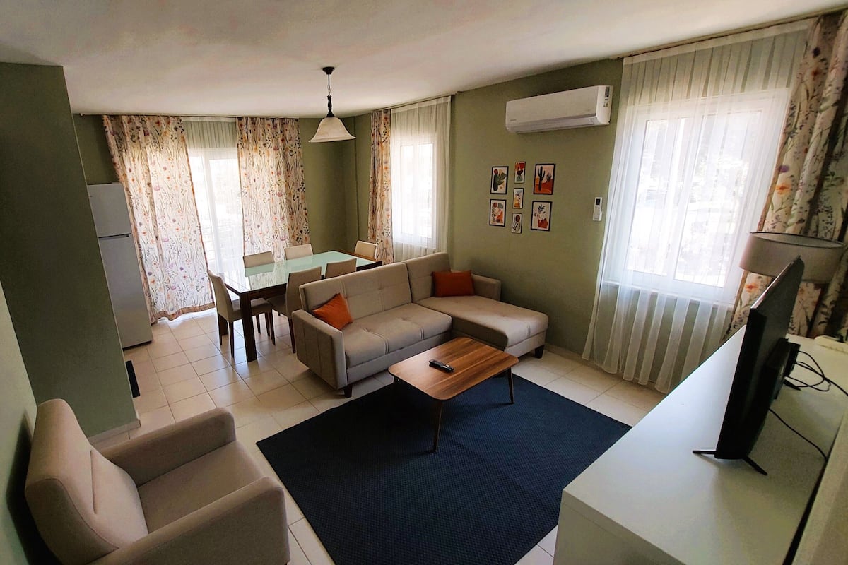 Een woonkamer met een bank, salontafel en een eettafel met stoelen. Er staat een tv op een dressoir, een koelkast, en een raam met gordijnen.