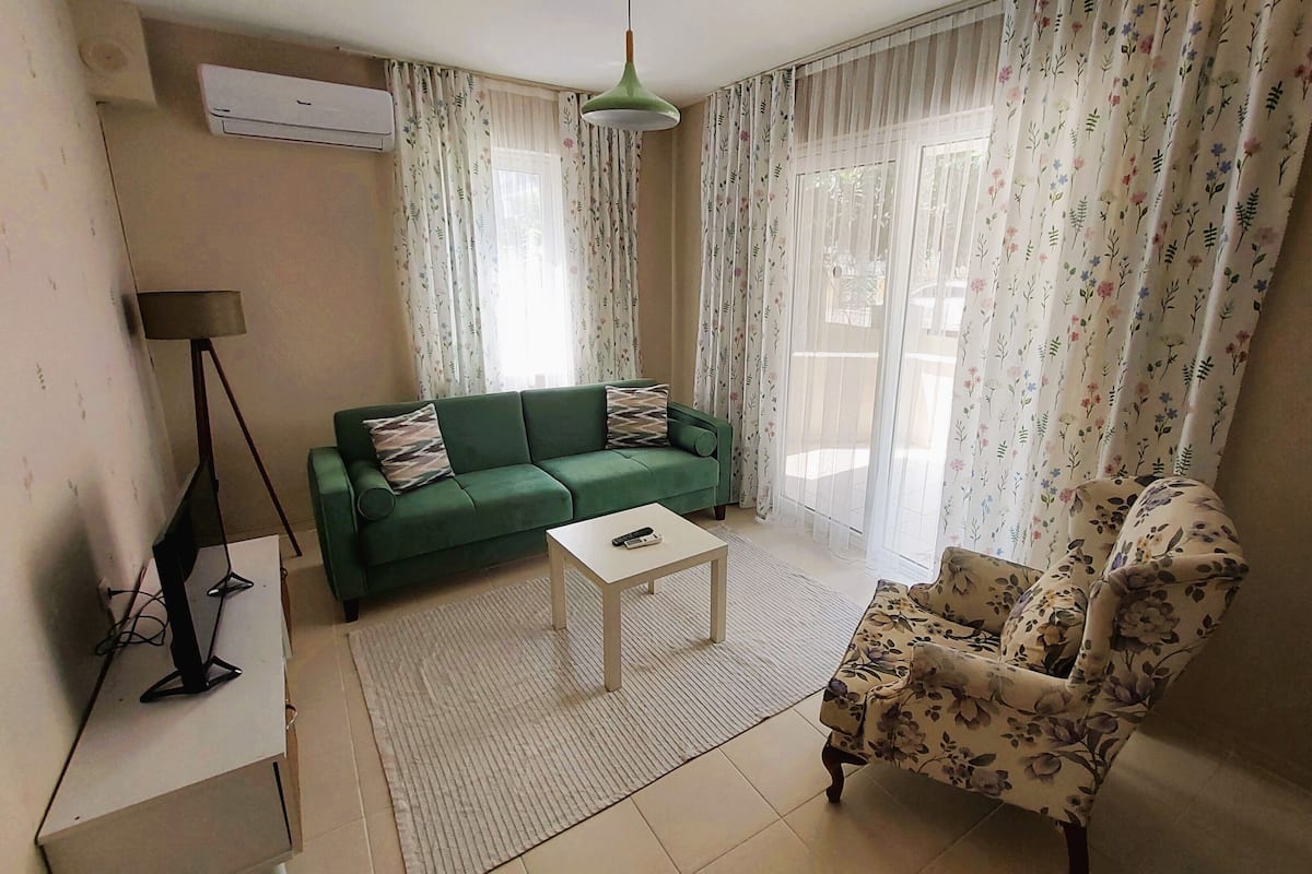 Een woonkamer met een groene bank, een bloemen fauteuil, een klein wit tafeltje en een vloerlamp.
