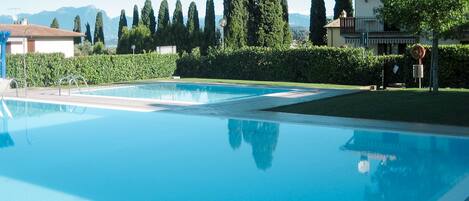 A clear blue swimming pool with metal ladders, surrounded by neatly trimmed hedges and tall trees.