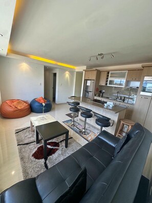 A modern living room with a black leather sofa, a coffee table, and two bean bags. A kitchen area with a bar and stools is visible in the background. - Furnished apartment with all the comforts to enjoy (mountain climate). (Valen)